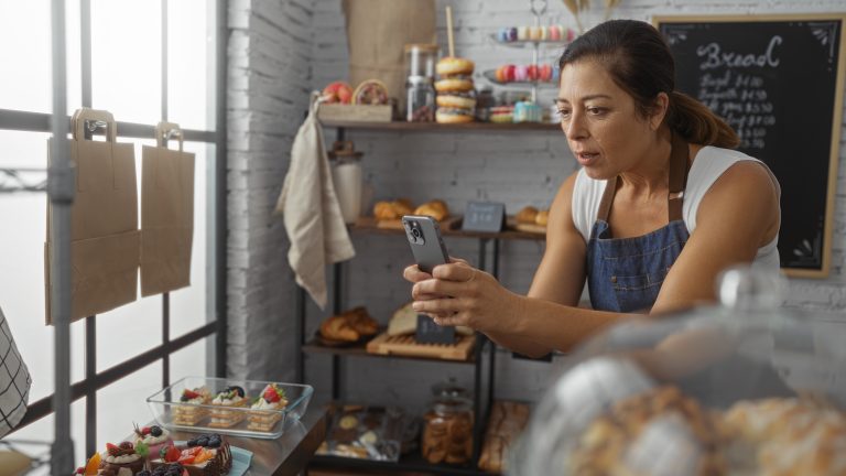 Small business owner using a smartphone to take a photo of pastries on a wooden countertop inside her shop, daylight from the window behind.