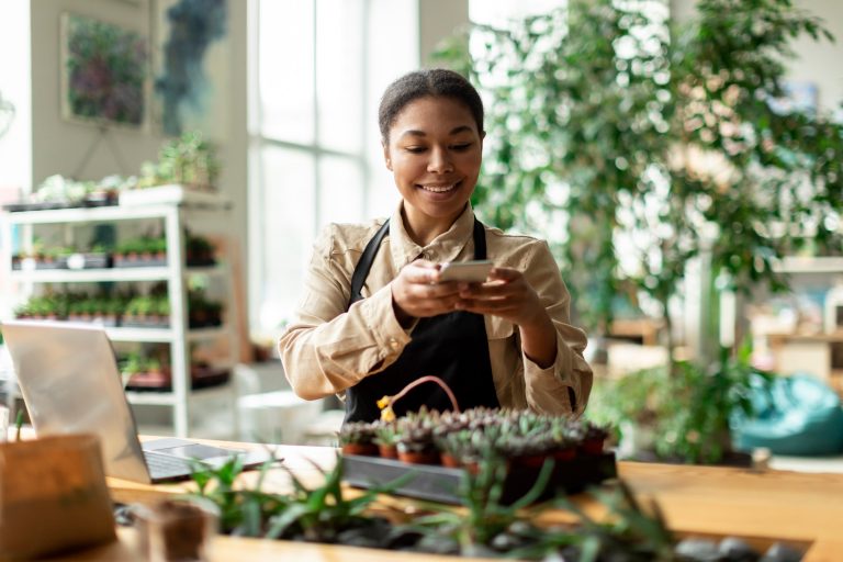 Plant shop owner in a work apron photographing a tray of her own succulents on a phone beside an open laptop