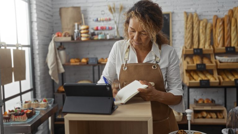 Bakery owner in an apron writing in a notebook beside a tablet at her shop counter