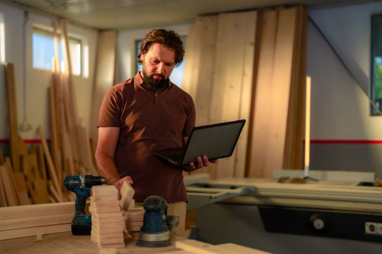 Woodworking shop owner standing at his workbench holding an open laptop and a cut wood block
