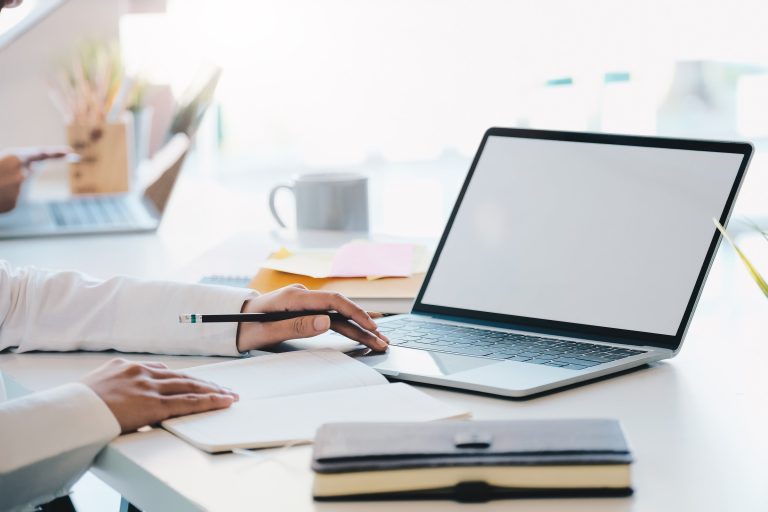 Business owner writing notes on a pad beside an open laptop on a bright office desk