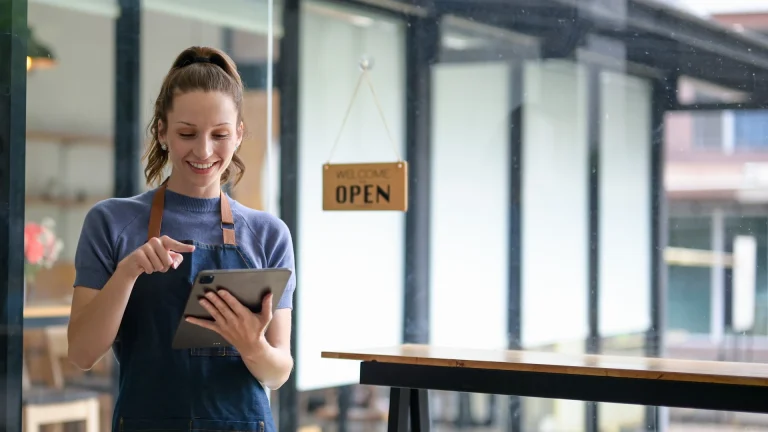 Smiling woman in an apron using a tablet beside a shop window with an Open sign