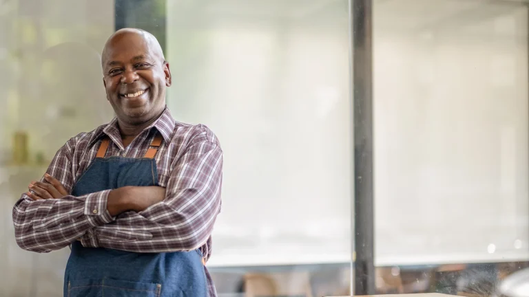 Smiling small business owner in a denim apron standing with arms crossed inside his shop