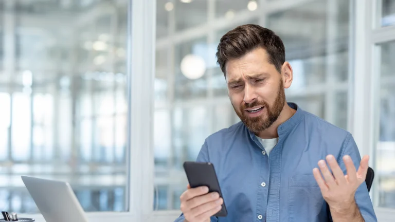 Frustrated man frowning at his smartphone and raising a hand in exasperation at his desk