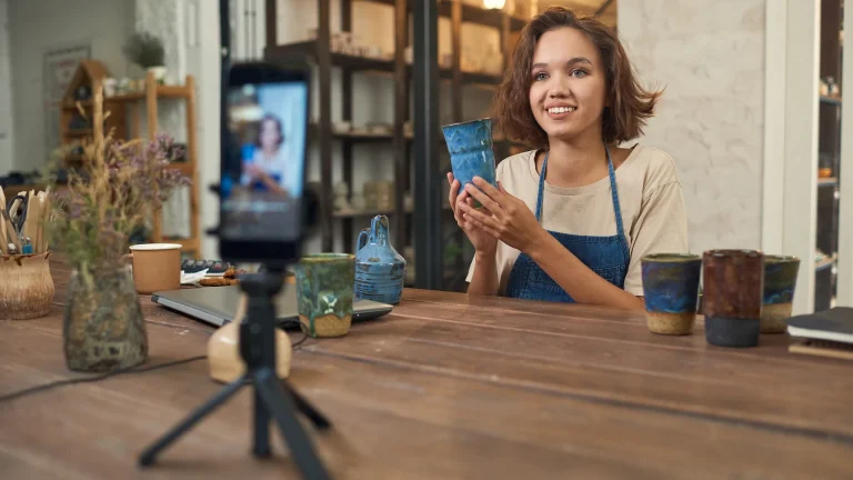 Woman in a blue apron holding a cup toward a smartphone on a tripod