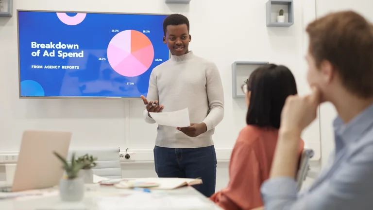 Man presenting a slide titled Breakdown of Ad Spend with a pie chart to two seated people