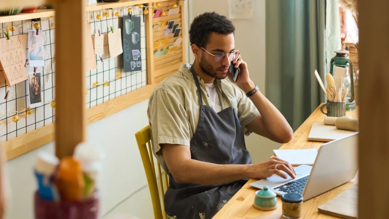 Man in an apron taking a phone call while using a laptop at a workbench