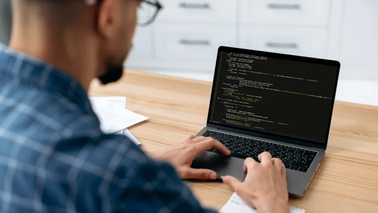 Over-the-shoulder view of a person typing on a laptop displaying code at a wooden desk