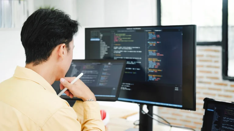 Person studying code on a large monitor and a smaller screen at a bright desk