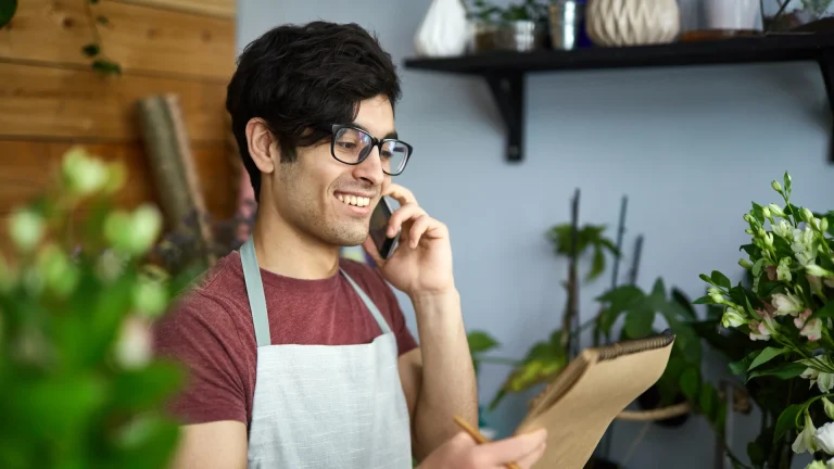 Smiling man in an apron holding a phone to his ear and a clipboard among flowers