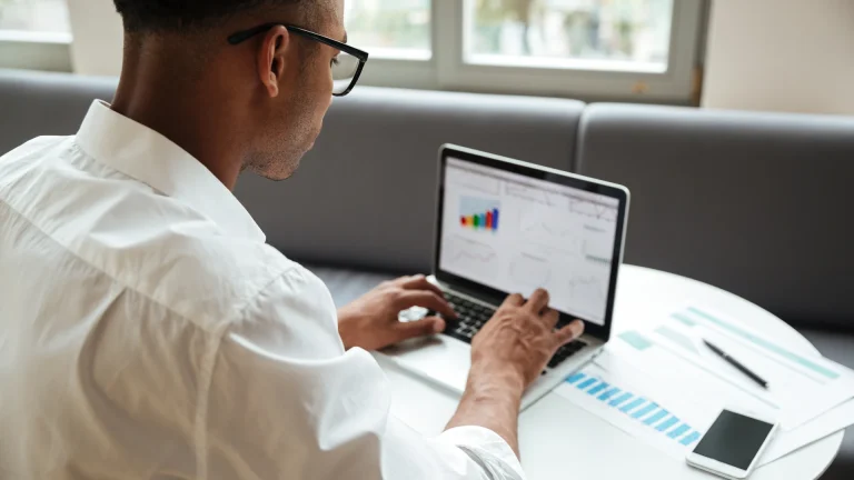 Man in glasses reviewing charts on a laptop beside printed charts and a phone
