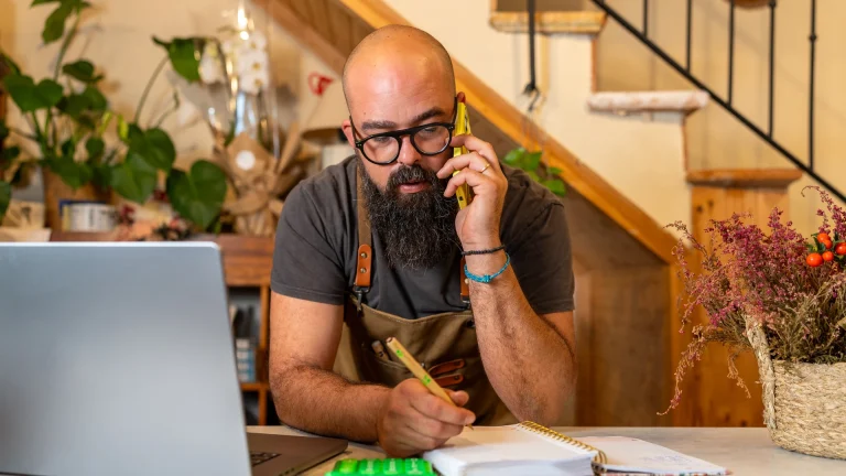 Bearded man in a work apron on a phone call at a shop counter, jotting notes beside a laptop