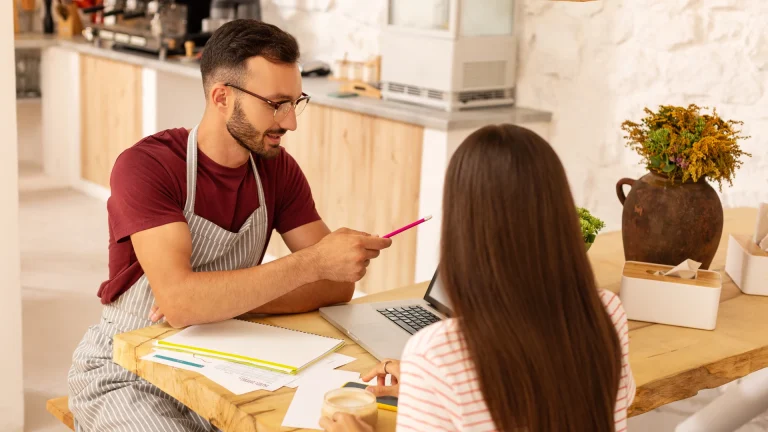 Man in a striped apron points a pencil while talking over papers with a woman at a laptop