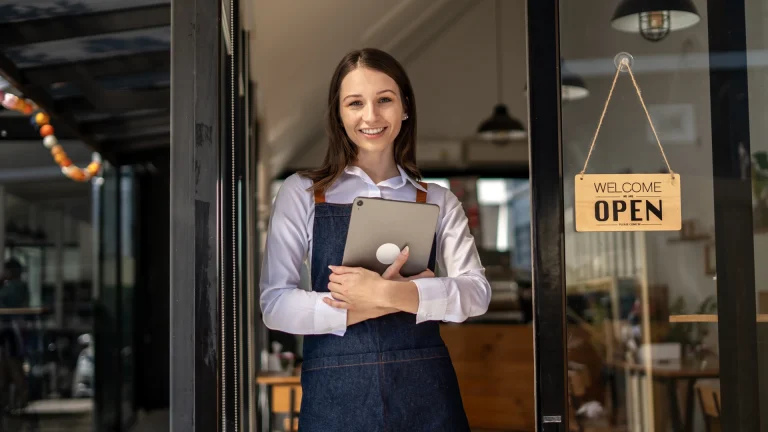 Smiling woman in a blue apron holding a tablet in a shop doorway beside an Open sign