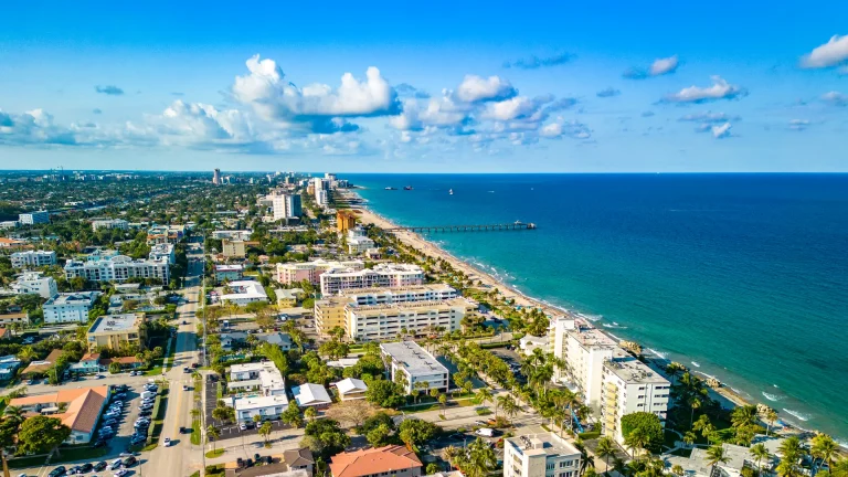Aerial view of coastal buildings, palm-lined streets, a sandy beach and a pier extending into the ocean
