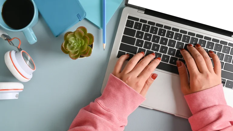 Overhead view of hands typing on a laptop beside a coffee mug, notebook, pencil and succulent