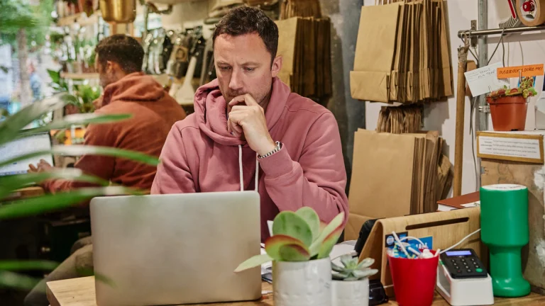 Man in a pink hoodie resting his chin on his hand while reading a laptop screen at a shop counter, with paper bags, a plant and a card reader nearby