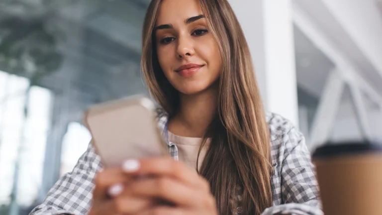 Woman in a gray gingham shirt looking down at a smartphone in her hands, with a paper coffee cup beside her