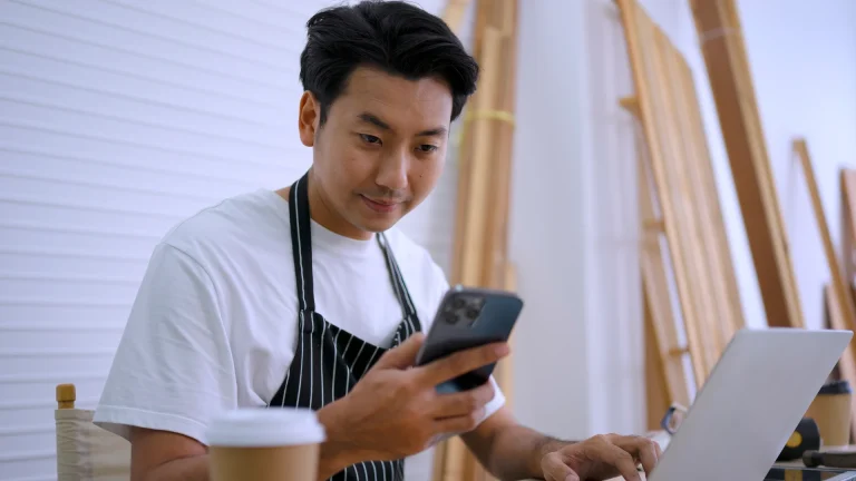Man in a striped apron checking a smartphone while typing on a laptop in a workshop