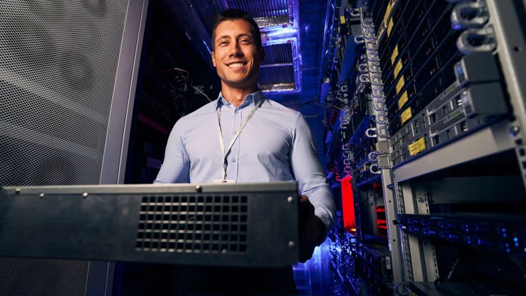 A smiling man in a light blue shirt and lanyard holds a server chassis in an aisle between lit server racks
