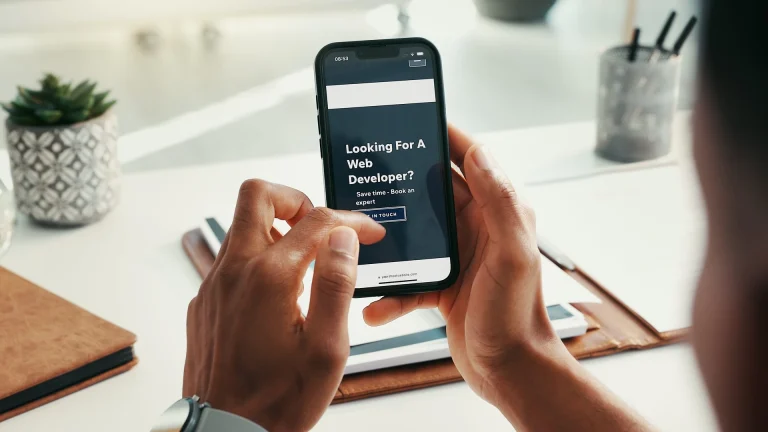 Two hands holding a smartphone that shows a dark web page headed Looking For A Web Developer, above a desk with notebooks, a succulent in a patterned pot and a pen holder
