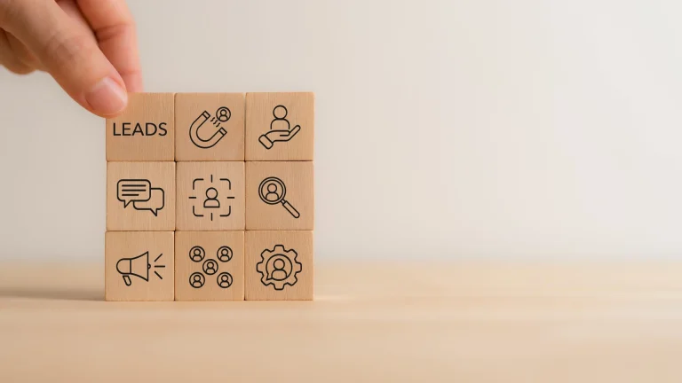 A hand placing a wooden block labeled LEADS on a three-by-three grid of blocks with line icons, including a magnet, a magnifying glass and a megaphone, against a pale wall