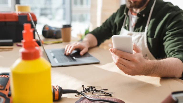 Man in a green shirt and apron holding a white smartphone in one hand and using a laptop with the other at a workbench with screws and power tools