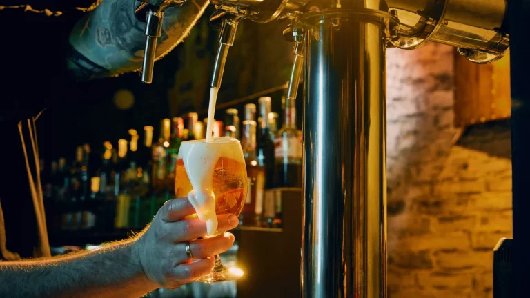 A hand holds a glass of foaming beer under a chrome bar tap, with bottles lined up on shelves behind