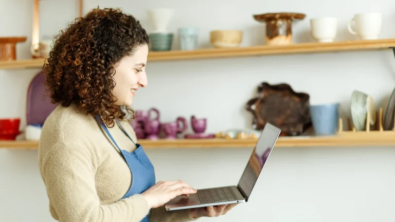 Woman in a blue apron standing in a pottery studio and typing on an open laptop, with shelves of ceramics behind her