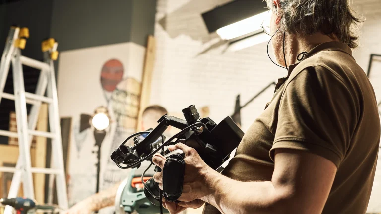 Camera operator in a brown polo shirt holding a professional video camera in a workshop, with a stepladder, a studio light and a second person partly visible in the background