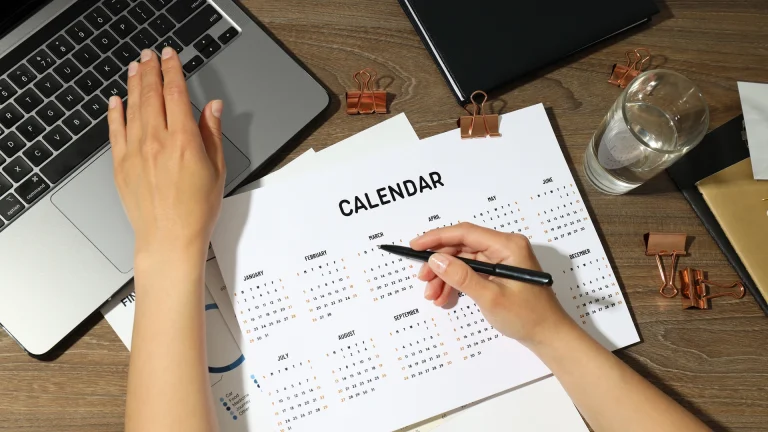 Overhead view of a person's hands on a wooden desk, one on a laptop and one holding a pen over a printed twelve-month calendar