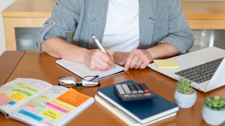 A person in a gray jacket writes in a notebook at a wooden desk beside an open laptop, a calculator, glasses and a planner covered in colored sticky notes