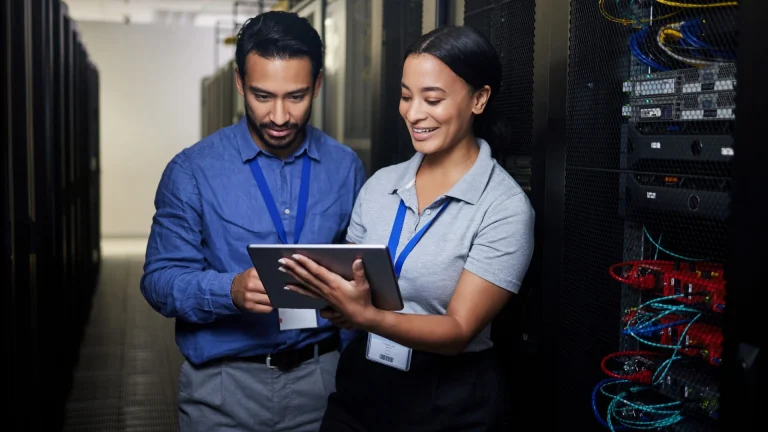 A man in a blue shirt and a woman in a gray polo, both wearing blue lanyards, look at a tablet in an aisle beside server racks with colored network cables