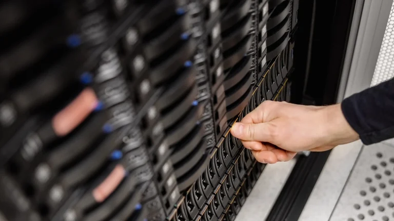 A hand gripping the handle of a drive tray in a rack of black server drive bays with blue latches, beside a perforated floor tile