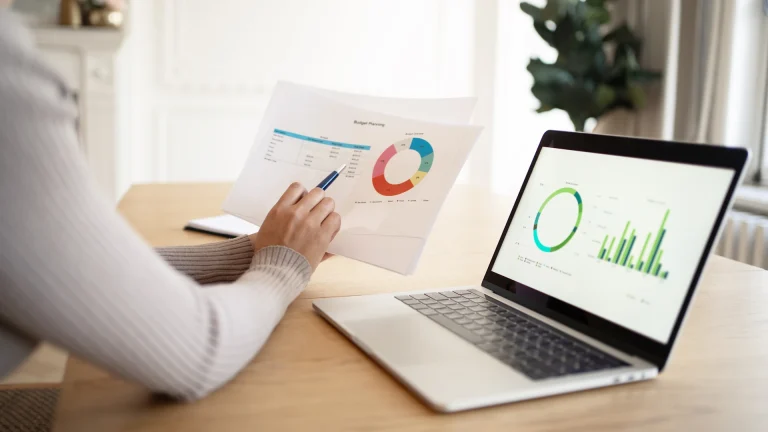 Hand pointing a pen at a printed page headed Budget Planning with a ring chart, beside an open laptop showing a ring chart and a bar chart on a wooden table