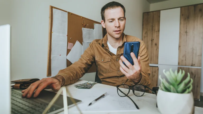 A man in a tan shirt at a desk looks at the smartphone in his hand while his other hand rests on a laptop keyboard